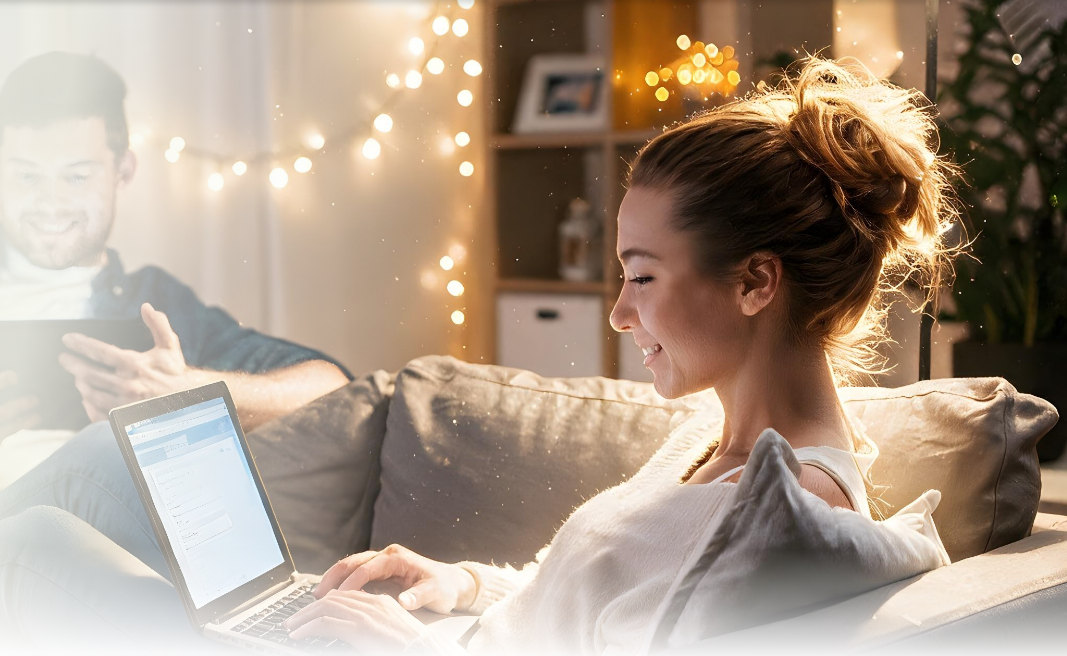 A woman sitting on a sofa using a laptop, with a man using a tablet in the background.